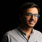 Close-up portrait of a young man wearing eyeglasses, smiling against a black background.